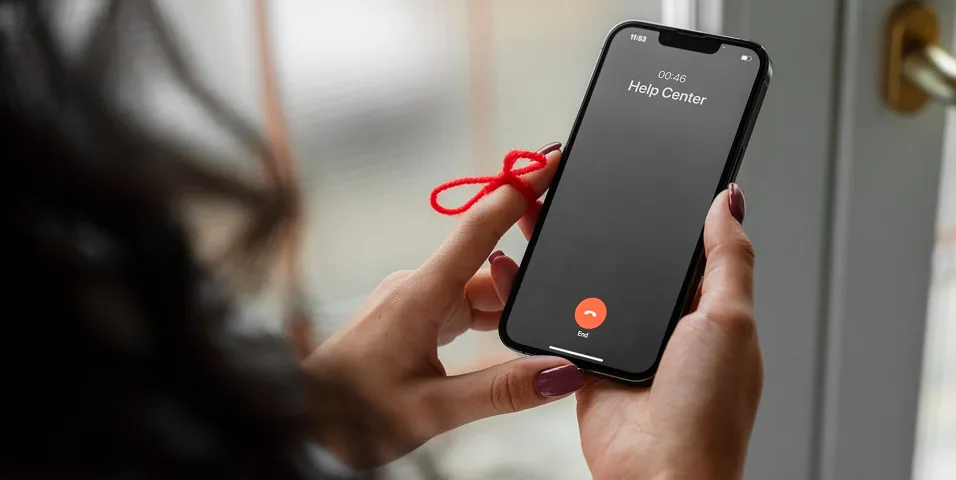 Pessoa segurando um smartphone mostrando uma chamada em andamento com a Central de Ajuda, com um cordão vermelho amarrado ao dedo.