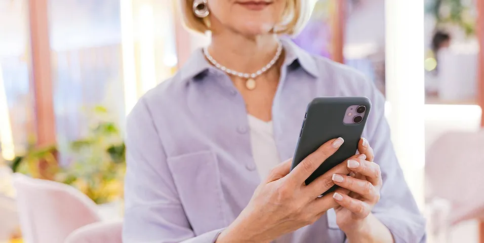 Mulher de meia-idade usando um colar e brincos, segurando e olhando para um smartphone dentro de casa.