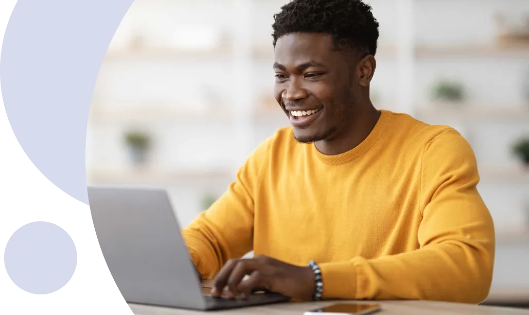 Hombre sonriente con un jersey amarillo escribiendo en un ordenador portátil en un escritorio con un smartphone cerca.