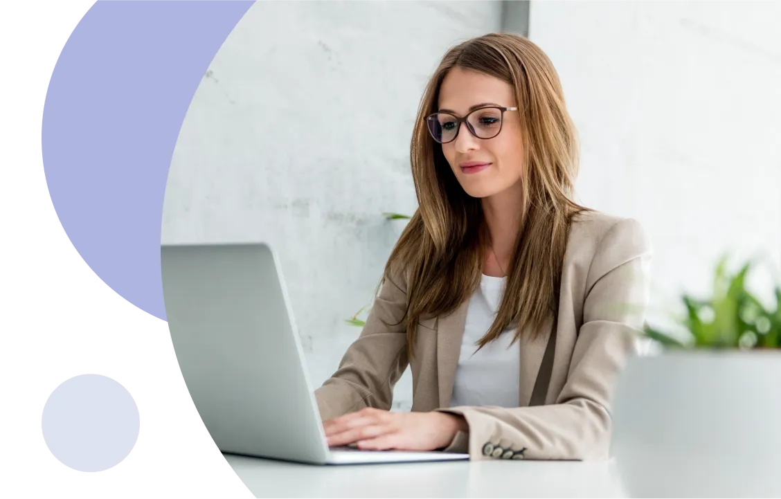 Mujer con gafas trabajando en un ordenador portátil en un escritorio blanco con una planta en primer plano.