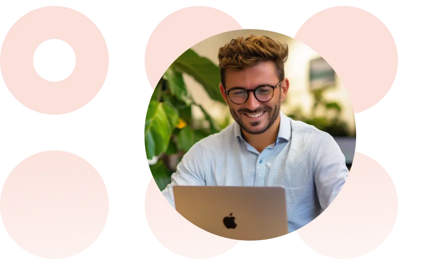 Homem sorridente com óculos trabalhando em um laptop em um ambiente interno bem iluminado, com plantas verdes ao fundo.