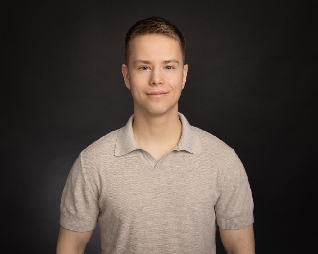 Nuori mies hymyilee neutraalia tummaa taustaa vasten, hänellä on vaalea lyhythihainen paita.
