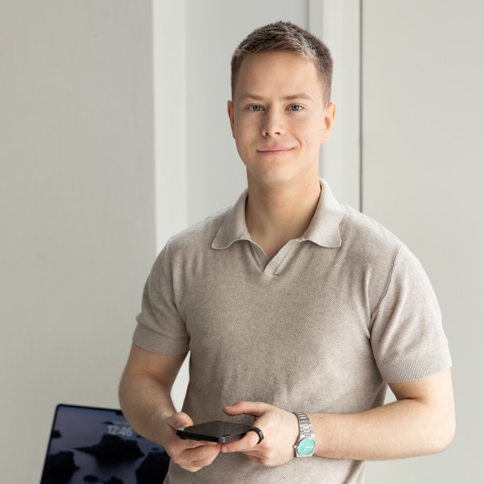 Nuori mies vaaleassa paidassa hymyilee ja pitelee puhelinta, taustalla näkyy kannettava tietokone.