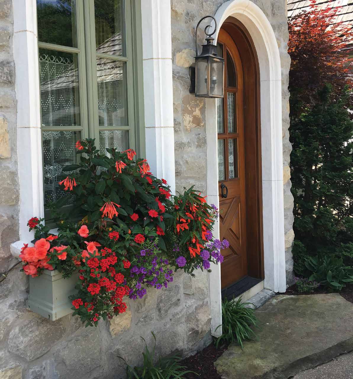 Stone house exterior with a wooden arched door and a window box filled with red and purple flowers.