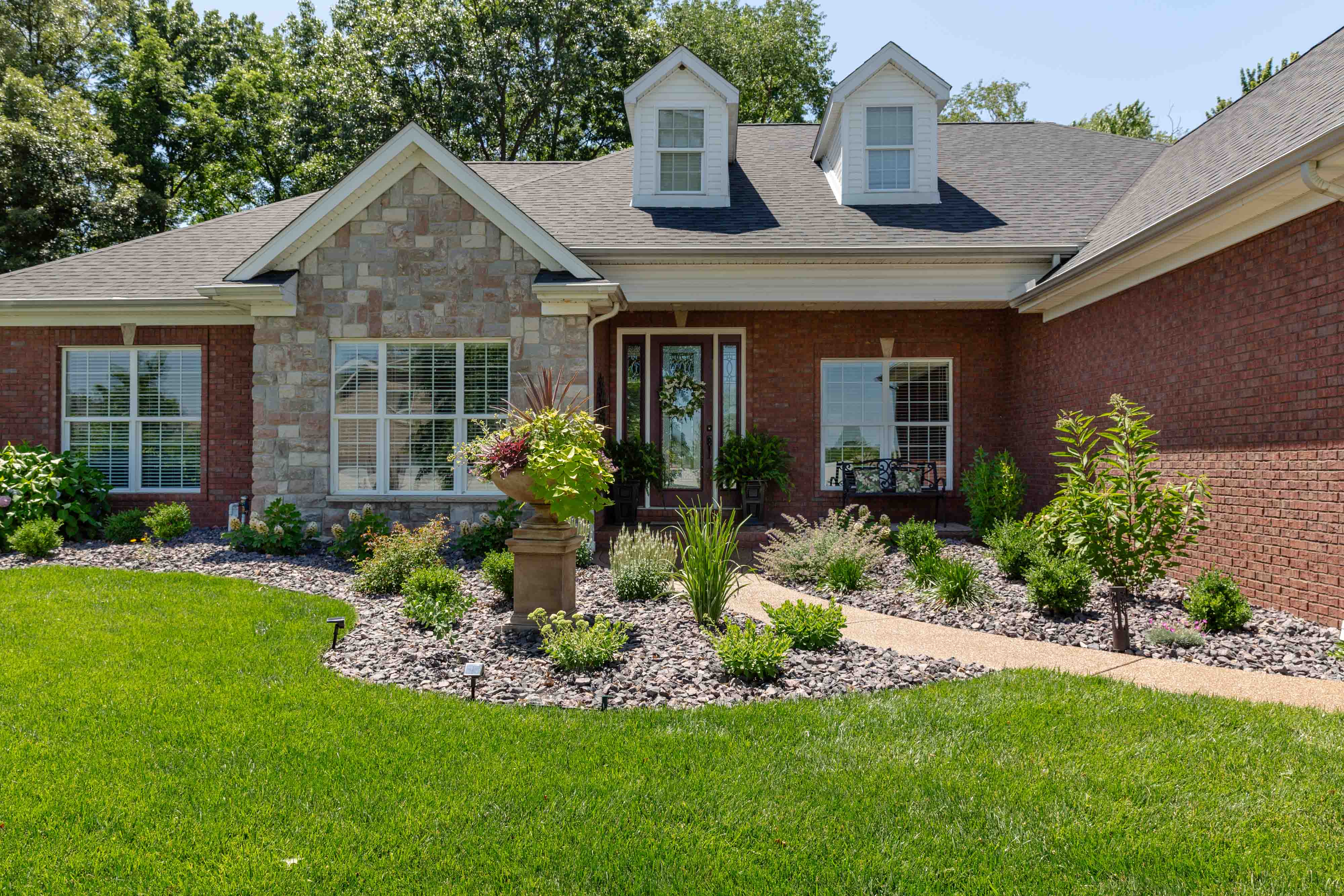 Front view of a brick house with stone accents, a decorated glass door, manicured garden beds, and a curved stone walkway through green lawn.