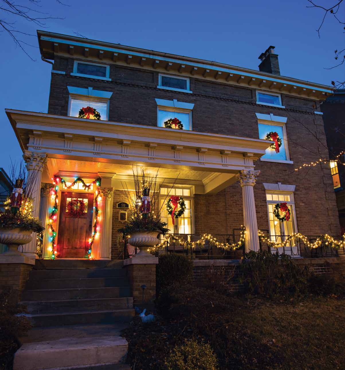 Two-story brick house decorated with Christmas wreaths, garlands, lights, and nutcracker figures at dusk.
