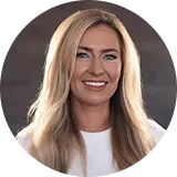 Smiling woman with long blonde hair wearing a white top against a blurred dark background.