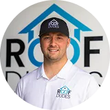 Smiling man wearing a black ROOF DUDES cap and white shirt with the ROOF DUDES logo in front of a matching logo backdrop.