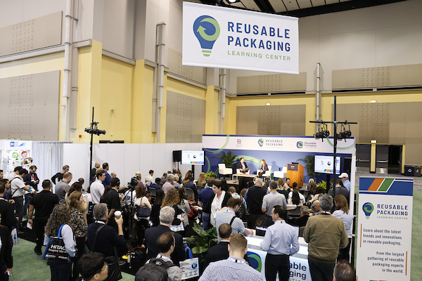 Crowd attending a presentation at the Reusable Packaging Learning Center with banners and a large screen panel.