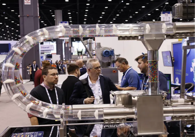 Group of men discussing around an industrial machine with a transparent curved conveyor carrying colorful small items at an exhibition.