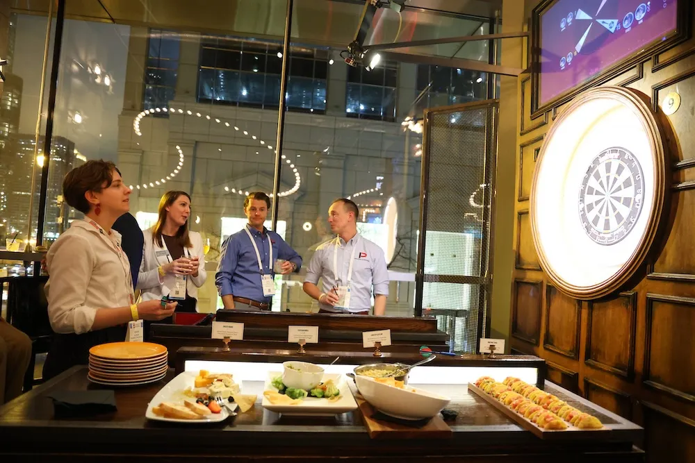 Four young adults standing and socializing near a dartboard in a dimly lit room with appetizers on a table in the foreground.