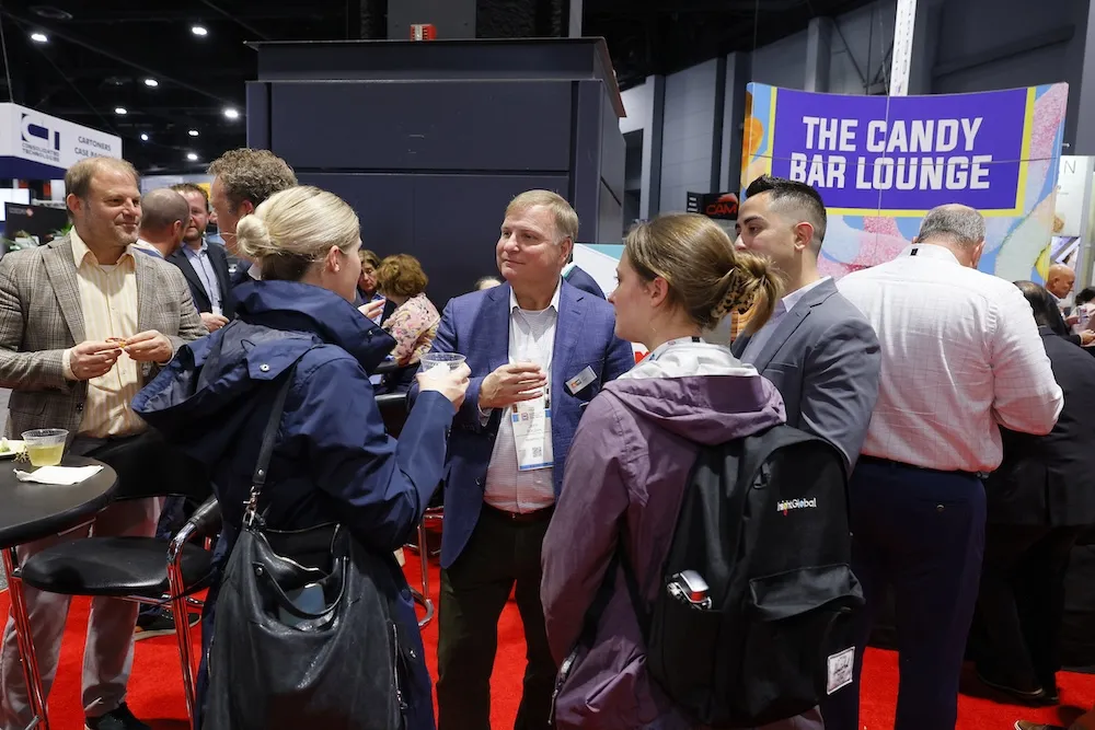 Group of people conversing and holding drinks near a booth with a sign that reads 'THE CANDY BAR LOUNGE' at an indoor event.