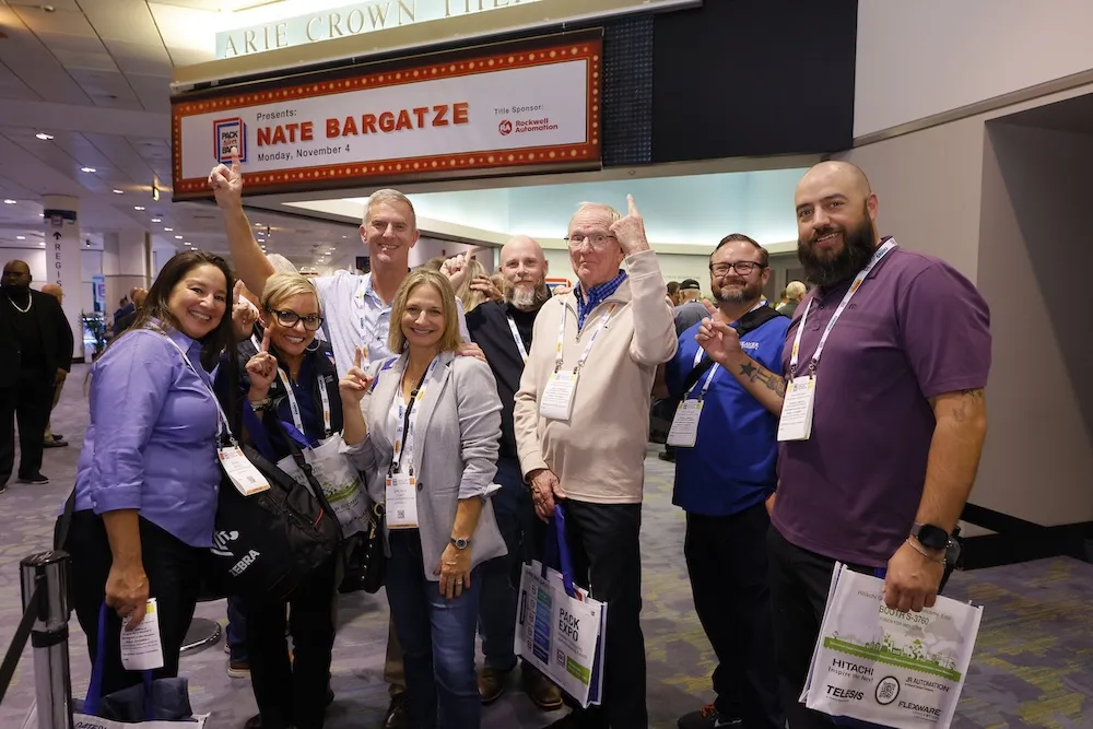 A group of smiling conference attendees posing and pointing upwards toward a sign for Nate Bargatze at the Arie Crown Theater.