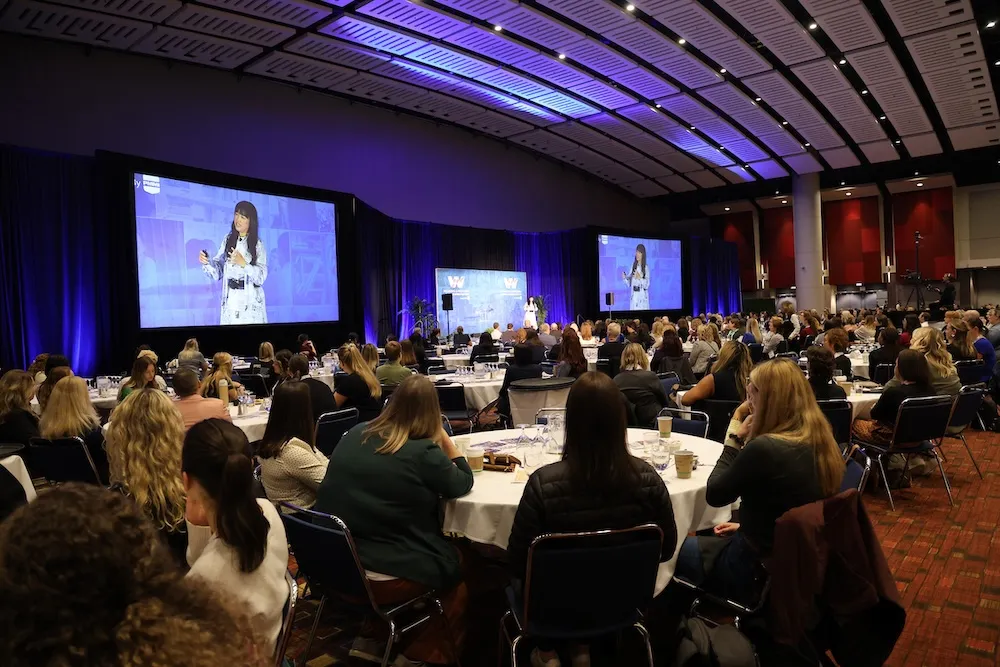 Large conference room filled with seated attendees watching a woman speaker presenting on stage with two large projection screens.