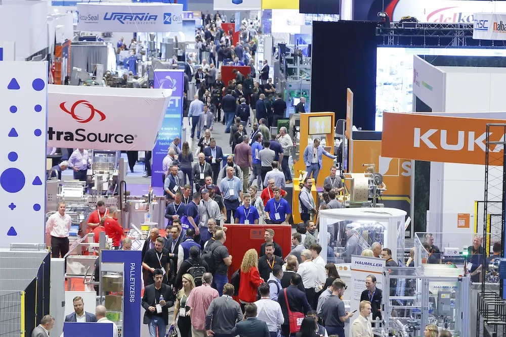 Crowded industrial trade show with people walking among booths and machinery displays.