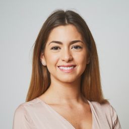 Smiling woman with long straight brown hair and light makeup wearing a pale pink blouse against a plain light background.