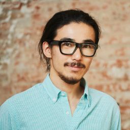 Two men, one holding a baby in yellow pajamas while the other gently touches the baby's hand, both smiling indoors.