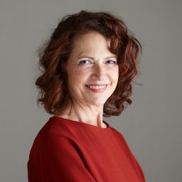 Smiling young woman with dark curly hair wearing a black blazer and heart-shaped pendant necklace.