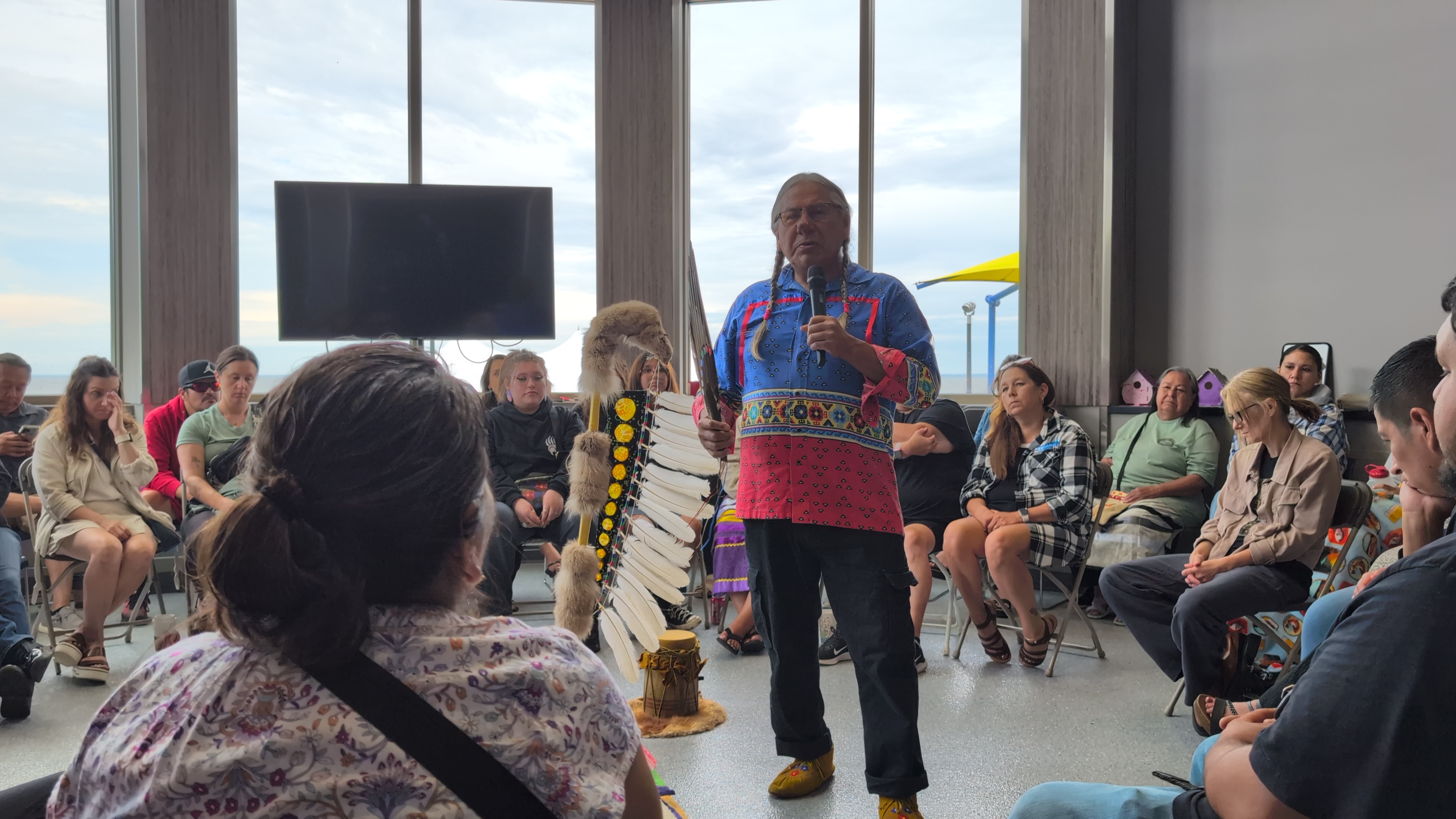 Man with braids wearing a colorful shirt speaks to a seated audience indoors near large windows.