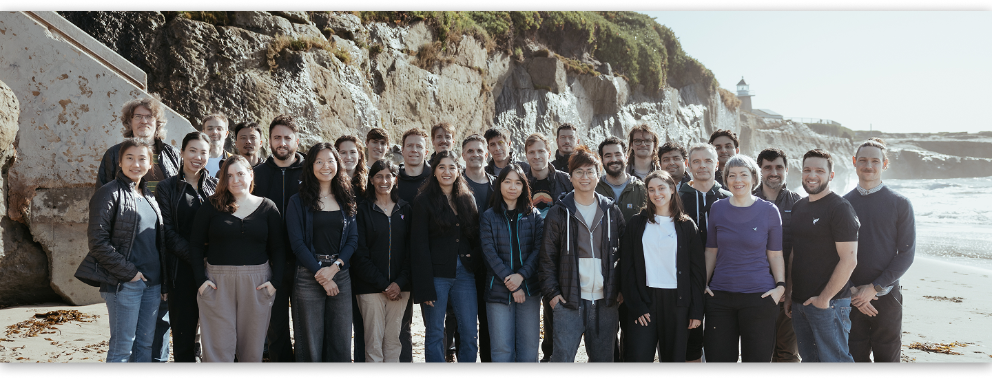 Group of 28 diverse people standing together on a sunny beach with rocky cliffs and a lighthouse in the background.