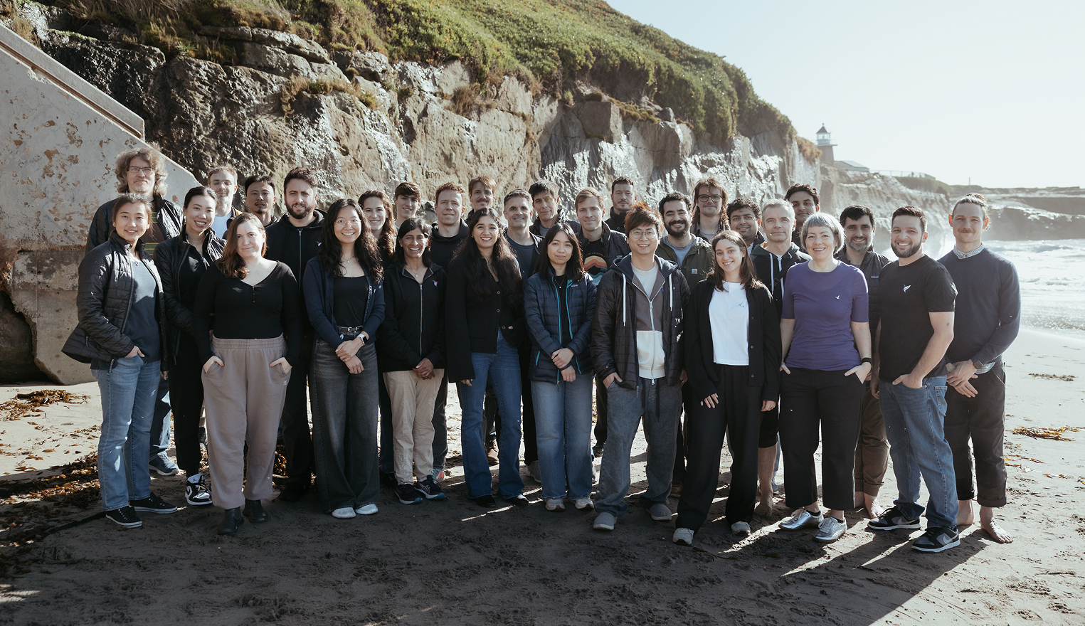 Group of diverse people standing together on a sandy beach with rocky cliffs and a lighthouse in the background.