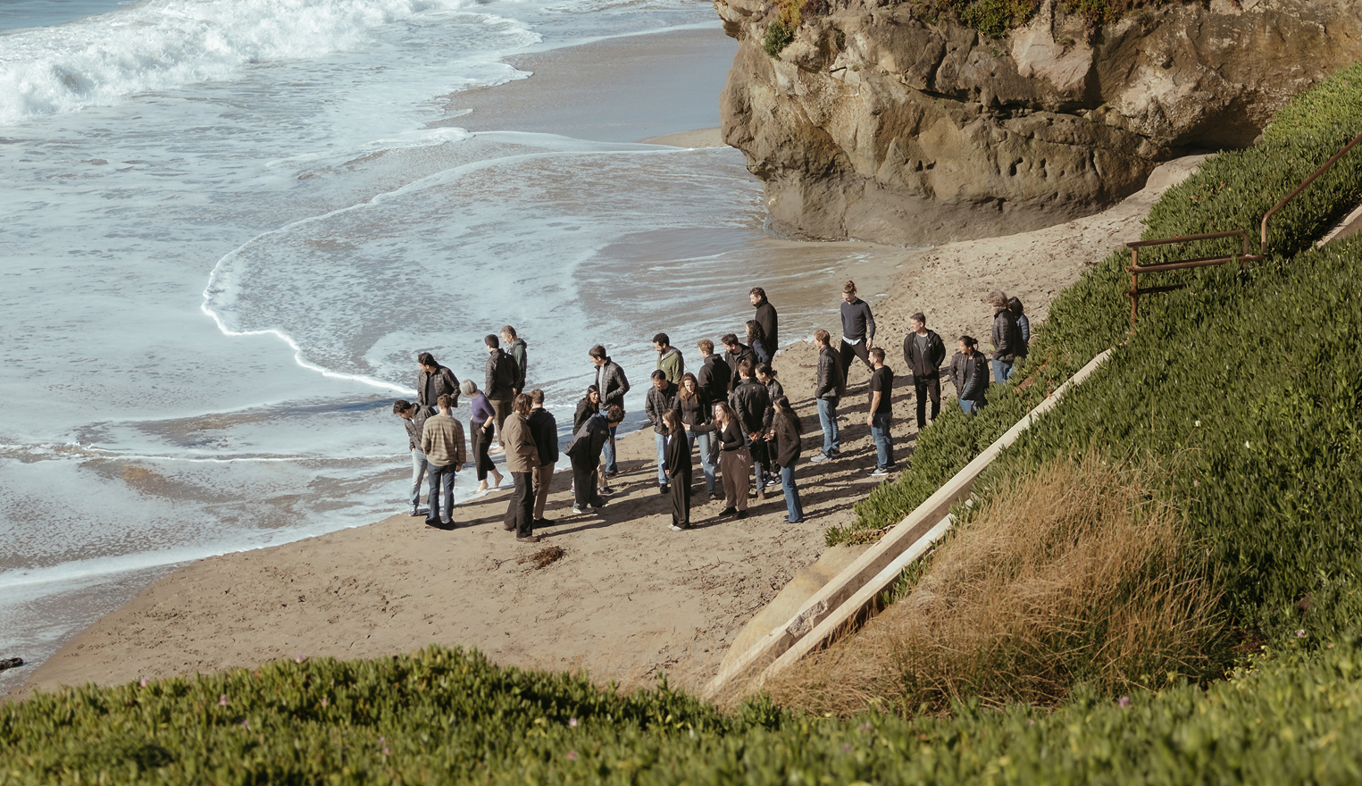 Group of people standing and talking on a sandy beach near ocean waves and rocky cliffs.