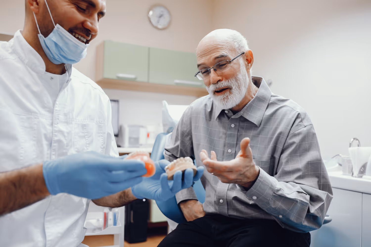 Dentist wearing gloves and a mask showing a dental model to an elderly man in a dental office.