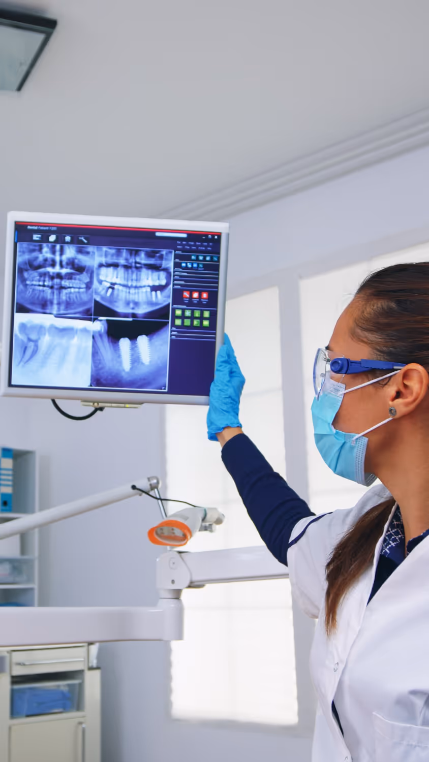 Female dentist wearing a mask and gloves examining dental X-rays on a monitor in a clinic.