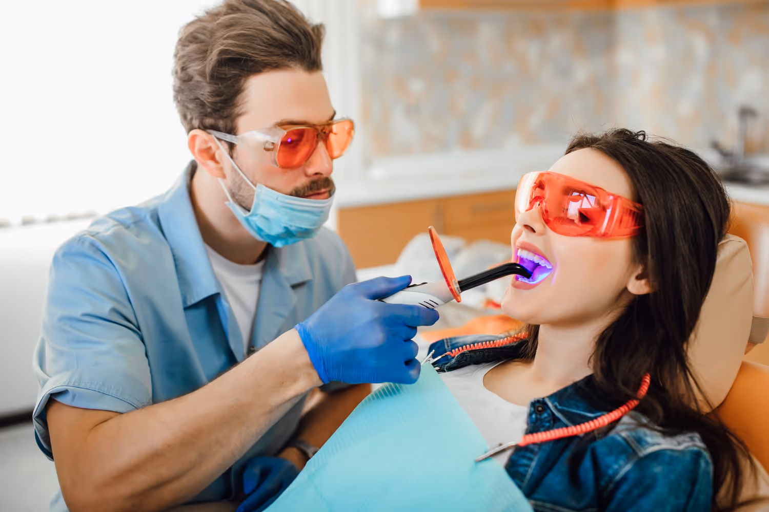 Dentist wearing protective glasses and mask uses a curing light device in the mouth of a reclining patient also wearing red protective glasses.