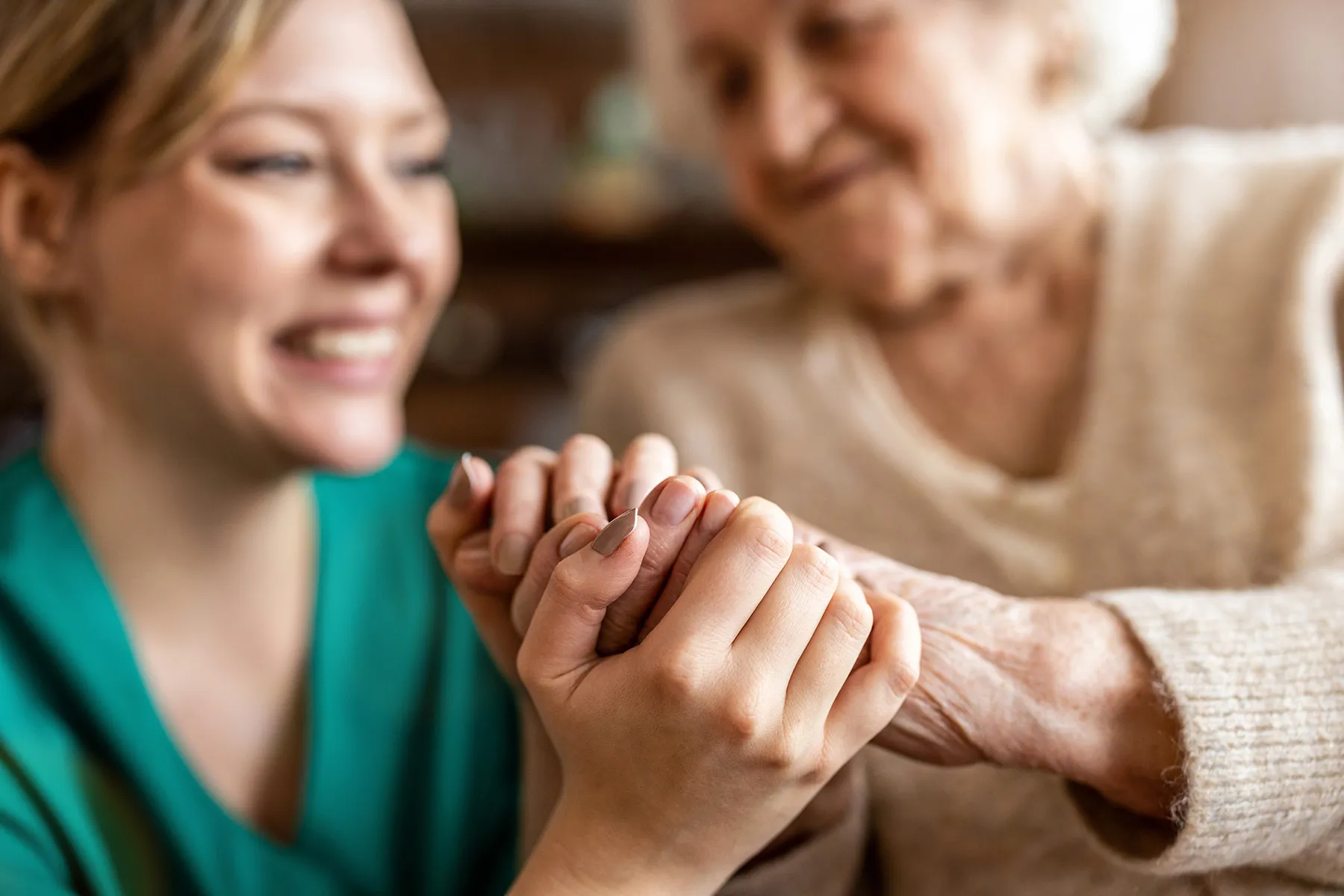 A smiling caregiver gently holds an elderly woman's hands, providing comfort and support.