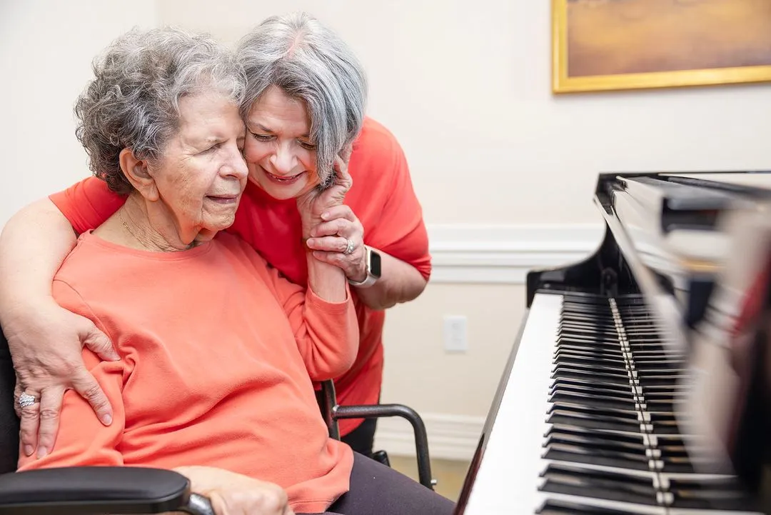Two elderly women in matching orange shirts share a warm embrace beside a piano.