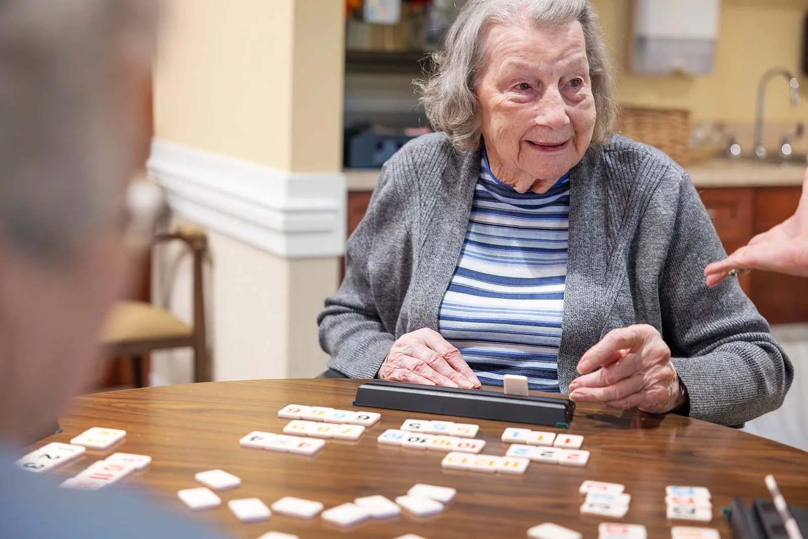 An elderly woman smiles while playing a tile game at a table.