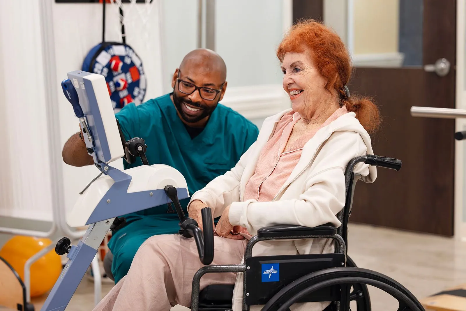 A man assists a smiling elderly woman in a wheelchair with using exercise equipment.
