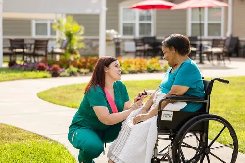 A caregiver kneels and talks to an elderly woman in a wheelchair outside on a sunny day.