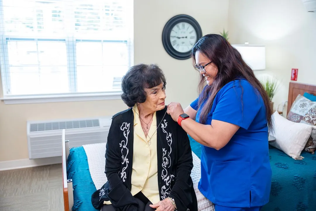 A healthcare worker helps an older woman sitting on a bed in a well-lit room.