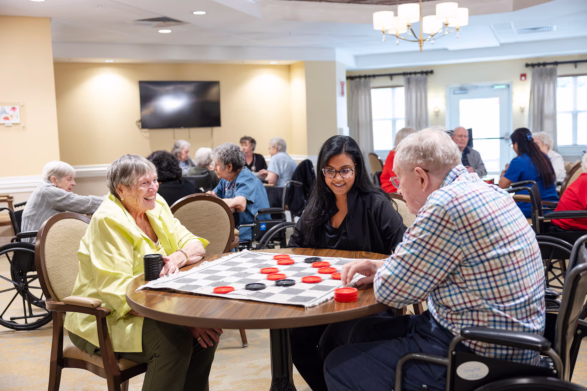 Older adults playing checkers at a table while a caregiver observes and smiles.