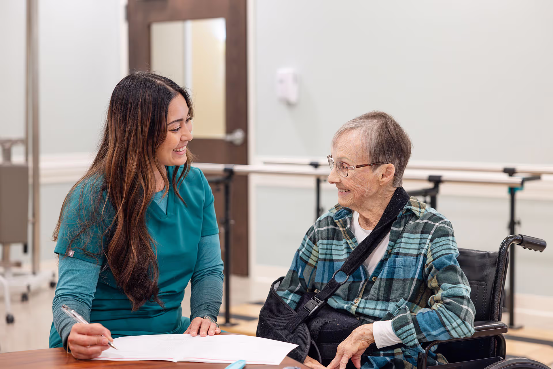 A healthcare worker in scrubs talks and smiles with an elderly person in a wheelchair.