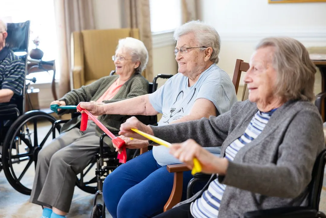 Three elderly women in wheelchairs exercising with resistance bands, smiling and seated in a well-lit room.