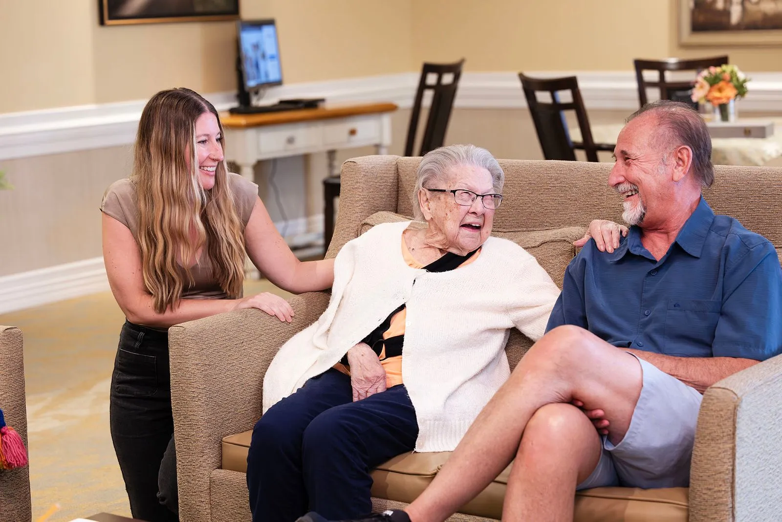 An elderly woman sits on a couch, smiling and talking with a man and a woman.