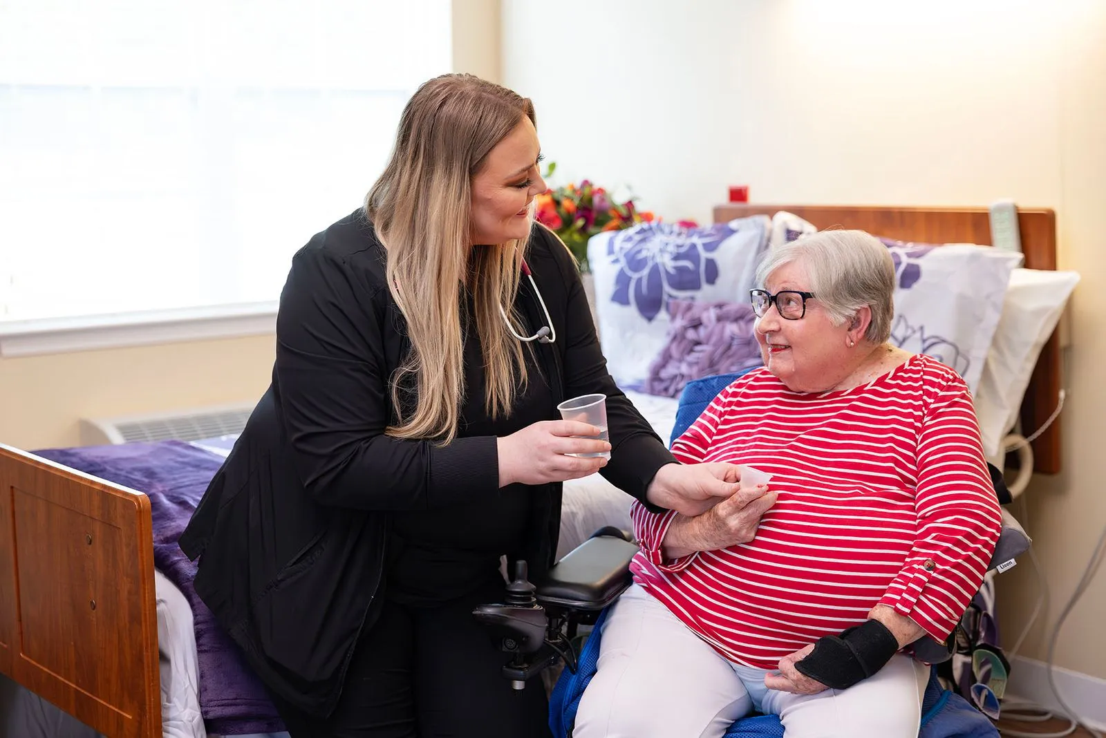A caregiver hands a cup to an elderly woman sitting on a bed, while they smile at each other.