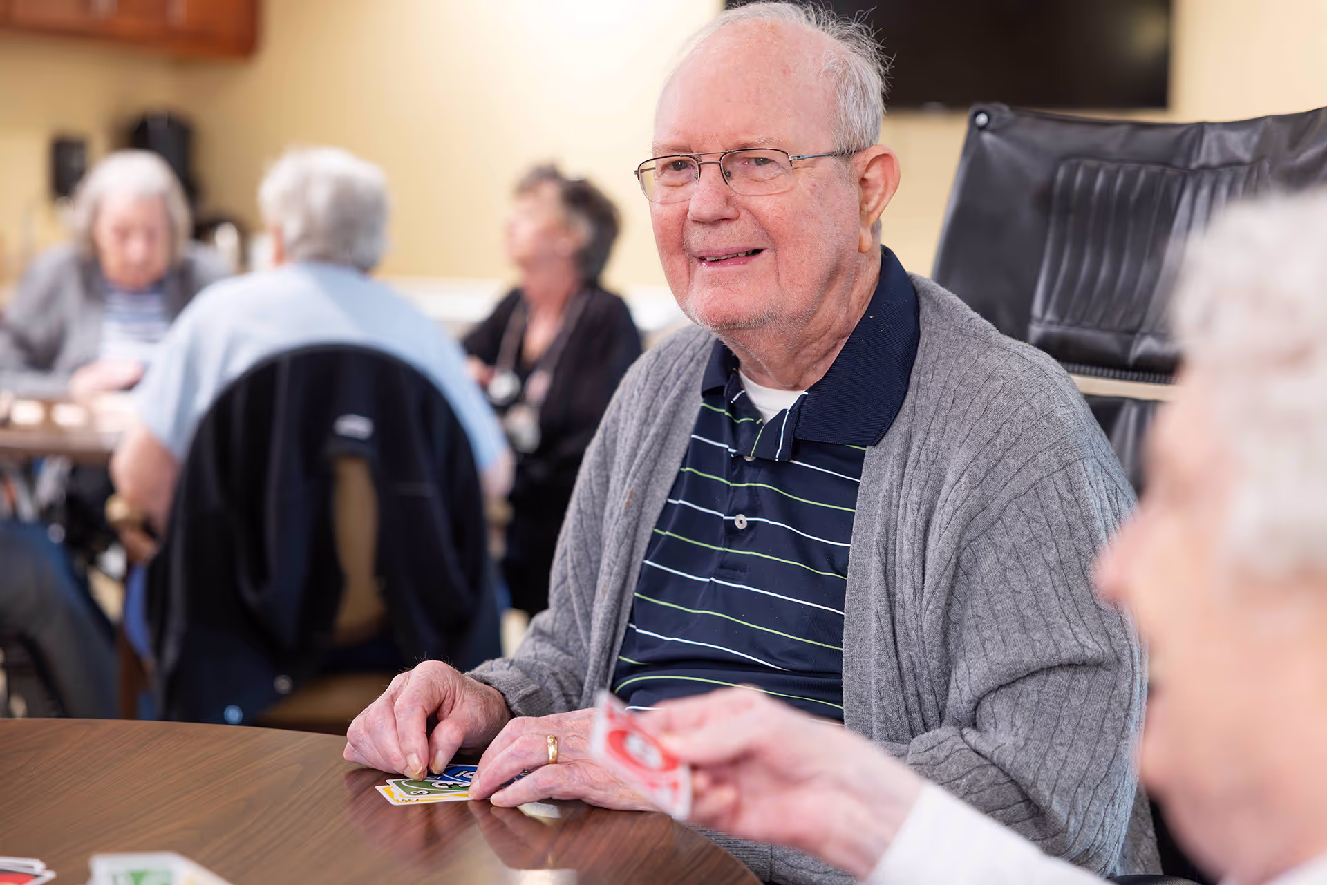 An elderly man smiles while playing cards with others at a table.
