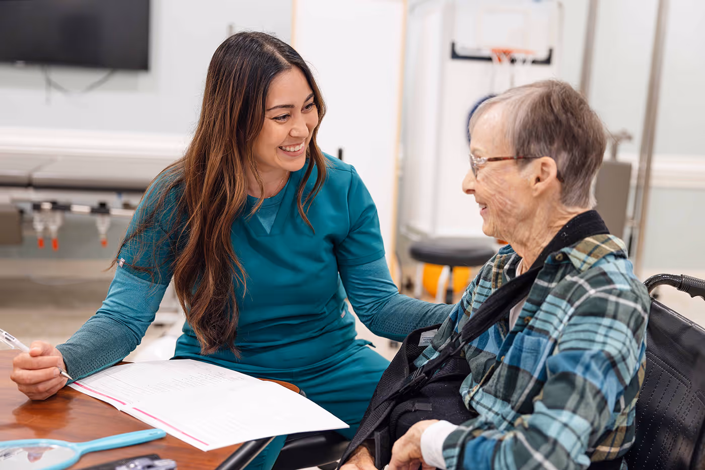 A healthcare worker smiles while talking to an older woman in a wheelchair.