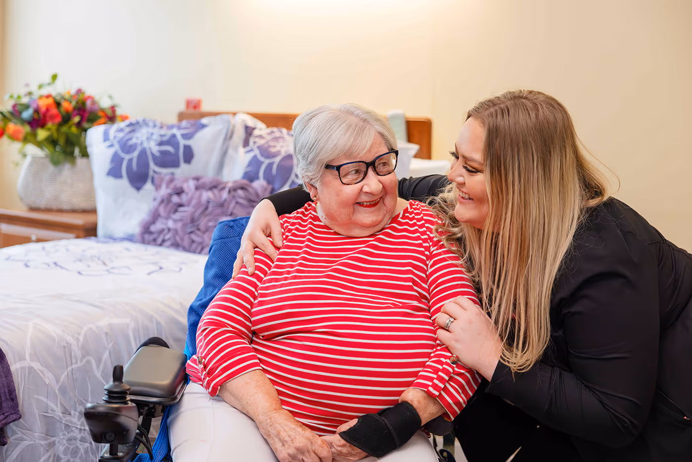 A woman leans in and smiles warmly at an elderly woman sitting in a wheelchair.