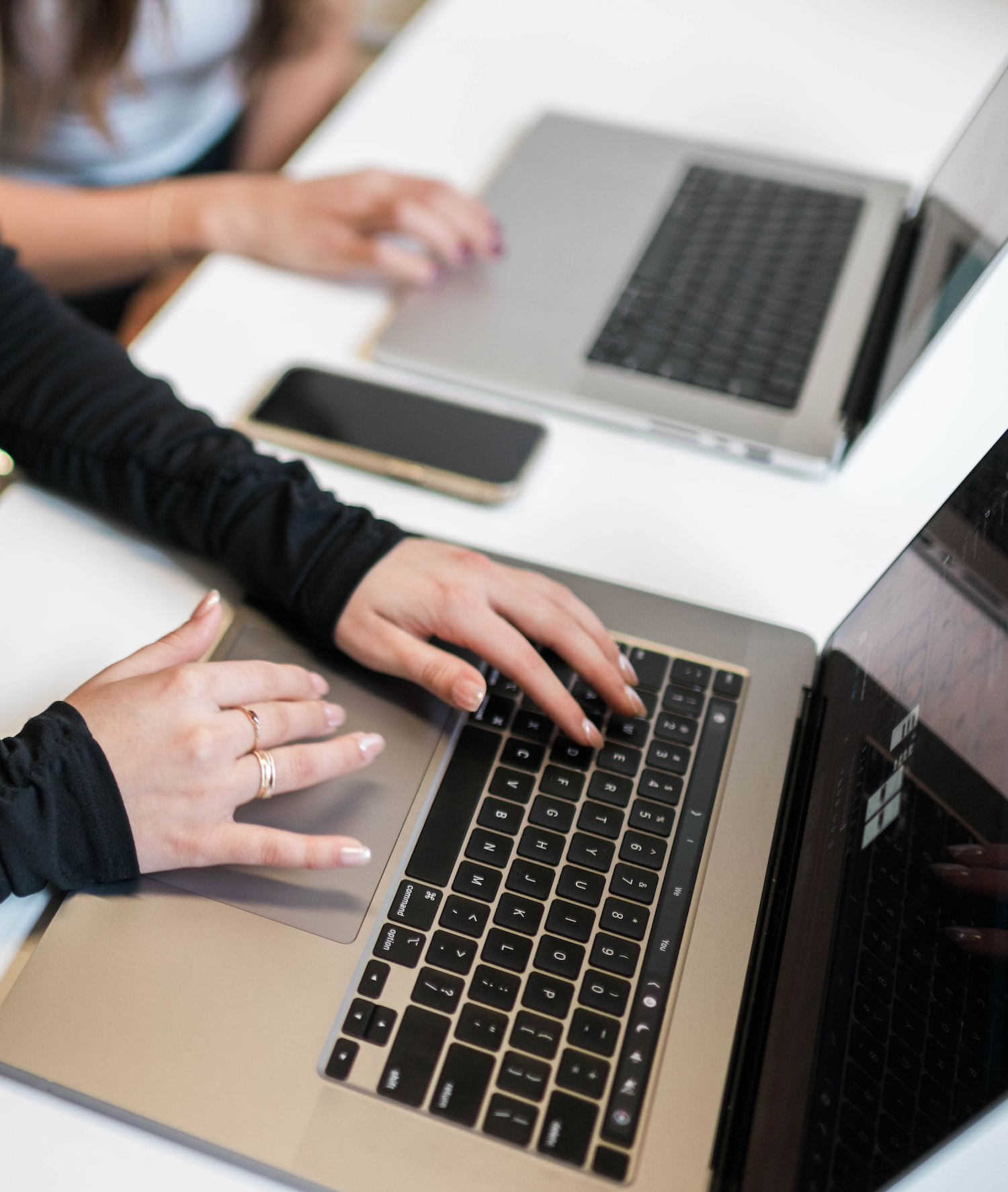 Two web designers working on laptops side by side on a white table with a smartphone between them.