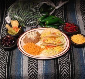 Plate with two tacos, rice, and beans, surrounded by bowls of chips, guacamole, black olives, shredded cheese, and whole peppers on a patterned cloth.