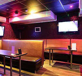 A cozy karaoke room with brown leather booths, two wall-mounted screens, and colorful ambient lighting.
