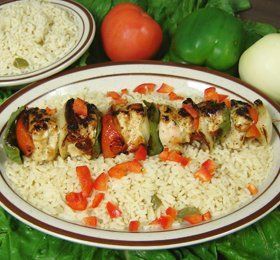 A plate of grilled chicken skewers with red bell peppers and onions on a bed of rice. Surrounded by fresh tomato and green pepper.