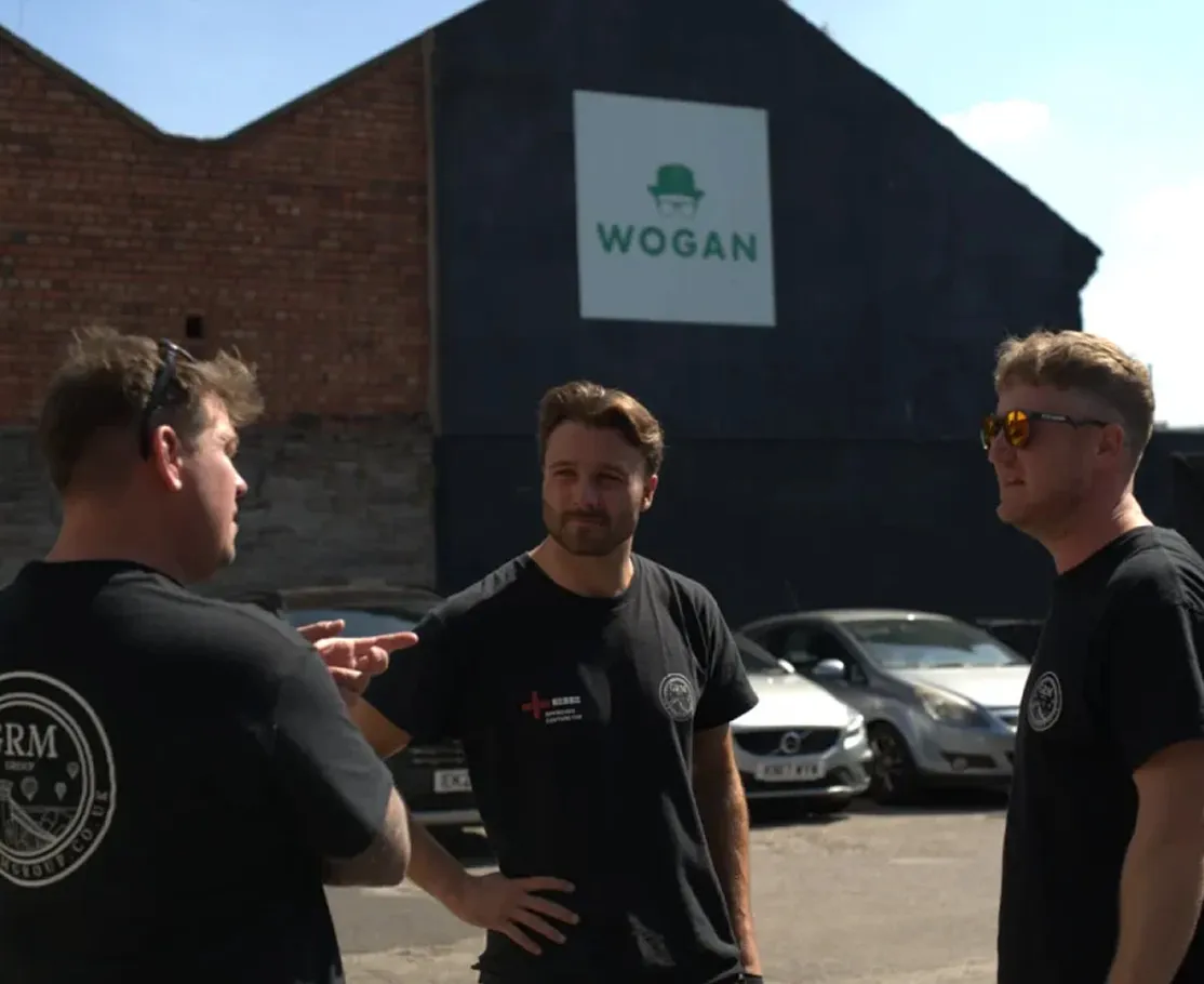 Three men wearing black t-shirts standing and talking in a parking lot with cars and a building bearing a WOGAN sign in the background.