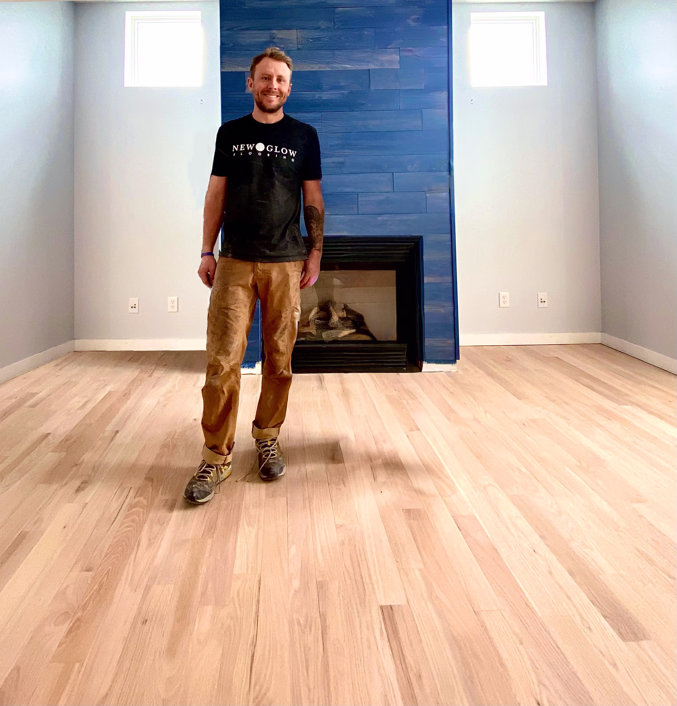 Man wearing a New Glow Flooring shirt standing on newly installed light wood flooring in a room with a blue-paneled fireplace.