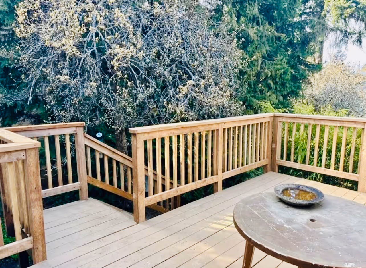 Wooden backyard deck with railing and stairs, a round table with a shallow dish, and trees in the background.